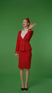 Canlı kırmızı üniformalı bir hostes yeşil bir geçmişe karşı neşeyle poz verir, profesyonellik ve dostluk yayar..