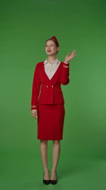 Kırmızı üniformalı neşeli bir uçuş görevlisi yeşil arka planda neşeyle el sallıyor. Ağır çekim. Kıyafeti profesyonelce ve şık, havayolu hizmetinin zarafetini ve çekiciliğini gösteriyor. Yavaş çekim.