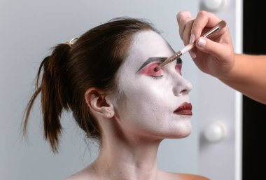Bir makyaj sanatçısı sahne makyajını beyaz yüz boyası ve kırmızı aksanlı yapar. Görünüşte çarpıcı bir teatral görünüş için çarpıcı kaşlar ve dudak şekillendirme ve yüz ifadeleri yer alıyor..