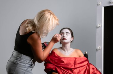 Bir makyöz, ayna önünde yumuşak ışıklandırma altında tiyatro için bir modele ustalıkla beyaz yüz boyası uygular..