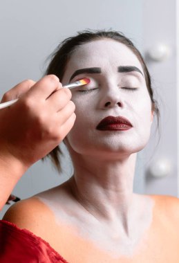 Yakından çekilen bir fotoğrafta beyaz boyalı bir yüze kırmızı göz farı süren bir el görülüyor. Dramatik makyajı vurgulanıyor..