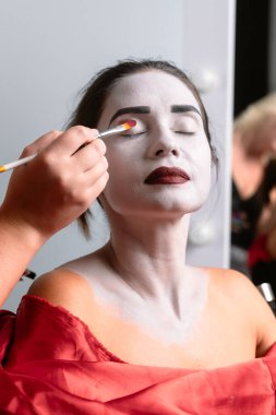 Yakından çekilen bir fotoğrafta beyaz boyalı bir yüze kırmızı göz farı süren bir el görülüyor. Dramatik makyajı vurgulanıyor..