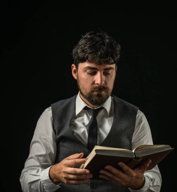 Görüntü, sakallı bir adamın resmi bir yelek ve kravat takıp kitap okuduğunu gösteriyor..