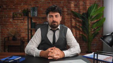 Resmi giyinmiş bir adam ahşap bir masada oturuyor, kırmızı tuğla bir duvar ve yeşil bir bitki arkaplanı var..