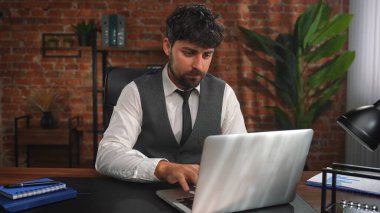 Yelek ve kravat giymiş odaklanmış bir adam masadaki dizüstü bilgisayarla çalışıyor. Bu da profesyonel bir ofis ortamını işaret ediyor..