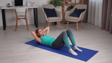 Genç bir kadın, içeride mavi bir minder üzerinde yoga egzersizleri yapıyor, esneklik ve gevşemeye odaklanıyor..