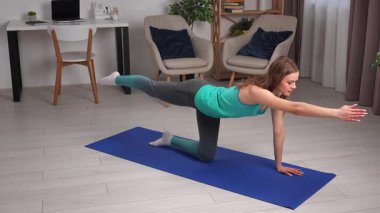 Genç bir kadın, içeride mavi bir minder üzerinde yoga egzersizleri yapıyor, esneklik ve gevşemeye odaklanıyor..