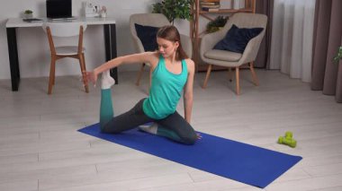 Genç bir kadın, içeride mavi bir minder üzerinde yoga egzersizleri yapıyor, esneklik ve gevşemeye odaklanıyor..