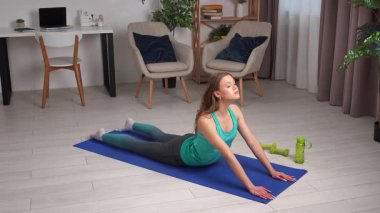 Genç bir kadın, içeride mavi bir minder üzerinde yoga egzersizleri yapıyor, esneklik ve gevşemeye odaklanıyor..