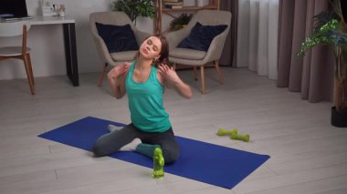 Genç bir kadın, içeride mavi bir minder üzerinde yoga egzersizleri yapıyor, esneklik ve gevşemeye odaklanıyor..