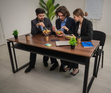Üç meslektaş bir masada sağlıklı bir öğle yemeğini paylaşıyor, bir şirket ortamında takım arkadaşlığı ve sağlığını vurguluyorlar..