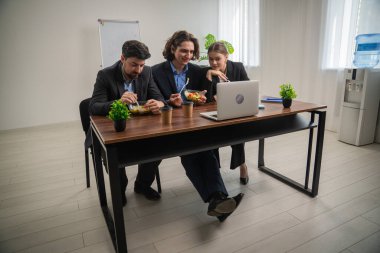 Üç meslektaş, modern ofis ortamında takım çalışması ve gevşemeyi vurgulamak için bir dizüstü bilgisayarla birlikte bir masada öğle yemeği arası veriyor..