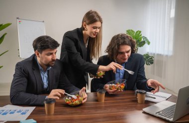 Bir grup profesyonel modern ofis ortamında proje detaylarını tartışırken sağlıklı bir öğle yemeği yiyorlar..