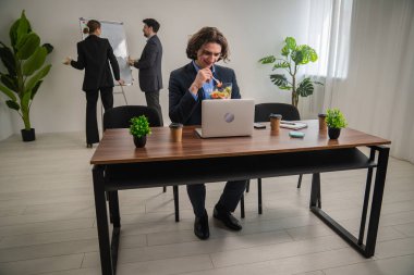 Meslektaşlar beyaz tahtada proje tartışırken diğeri masada sağlıklı bir yemeğin tadını çıkarıyor..