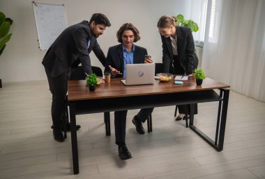 Bir grup profesyonel, modern ofis alanındaki bir toplantı sırasında dizüstü bilgisayarın etrafında bir tartışmaya girişiyor..