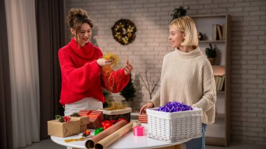 Şen süveter giymiş iki kadın, Noel ruhunu kucaklayan, sıcak dekore edilmiş bir salonda el yapımı hediyeleri neşeyle paketliyor..