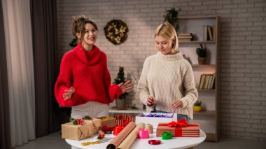 Şen süveter giymiş iki kadın, Noel ruhunu kucaklayan, sıcak dekore edilmiş bir salonda el yapımı hediyeleri neşeyle paketliyor..