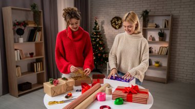 Şen süveter giymiş iki kadın, Noel ruhunu kucaklayan, sıcak dekore edilmiş bir salonda el yapımı hediyeleri neşeyle paketliyor..