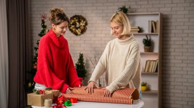 Sıcak dekore edilmiş bir oturma odasında, iki kız arkadaş heyecanla ev yapımı hediyeleri birlikte paketliyor. Şen süveter giyip tatil sezonunu kutlarken dostluk ruhunu kucaklıyorlar..