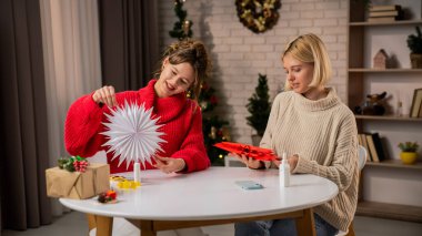 Kırmızı ve beyaz kazaklı iki kadın sıcak dekore edilmiş bir oturma odasında hediyeleri paketlerken kaliteli zaman geçirmekten hoşlanırlar. Noel süslemeleri neşe ve neşeyi paylaştıkça şenlikli bir ortam yaratır..