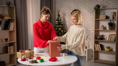 Sıcak bir oturma odasında, şenlikli kazaklı iki kız arkadaş ev yapımı hediyeleri dikkatlice paketliyor. Kış mevsiminin sıcaklığı onların neşeli dekorlarına ve tatil ruhlarına yansıyor..