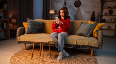 Genç bir kadın kanepede oturur ve akıllı bir telefona bakar. Dişi, şenlikli bir Noel kırmızı kazağı giyiyor. Ilık ışık odayı aydınlatır, sıcak kış atmosferini güçlendirir..