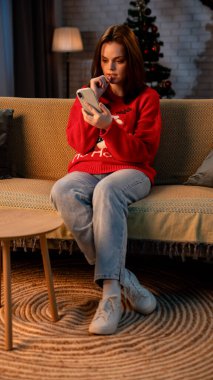 Genç bir kadın kanepede oturur ve akıllı bir telefona bakar. Dişi, şenlikli bir Noel kırmızı kazağı giyiyor. Ilık ışık odayı aydınlatır, sıcak kış atmosferini güçlendirir..