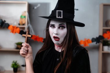 Vampir makyajı yapmış cadı bir kadın ve cadı şapkası Cadılar Bayramı için dekore edilmiş bir odada ahşap bir haç tutuyor. Korku filmleri, partiler ve gotik temalar için mükemmel..