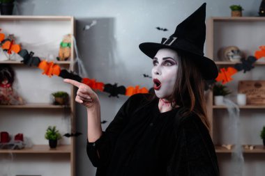 Vampir makyajlı ve siyah cadı şapkalı bir kadın Cadılar Bayramı için dekore edilmiş bir odada dururken parmağını yana doğru gösteriyor. Ürkütücü, neşeli ya da fantezi temalar için mükemmel..