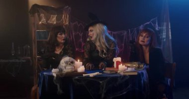 Witches order food on their smartphones and receive delivery from a ghost. Three women in witch costumes gather around a table with candles and a skull for a Halloween scene.