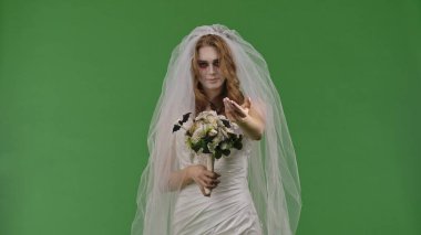 A woman dressed as a dead bride in a white wedding dress and veil makes a come here hand gesture. The ghostly bride stands against a green chroma key backdrop for Halloween or spooky projects.