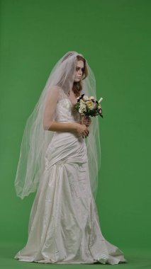 A woman dressed as a dead bride in a white wedding dress and veil holds a bouquet of flowers. The ghostly bride stands against a green chroma key backdrop for Halloween or spooky projects.
