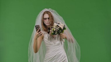 A woman dressed as a dead bride, in a white wedding dress and veil, browses information on a smartphone. The ghostly bride stands against a green chroma key backdrop for Halloween or spooky projects.