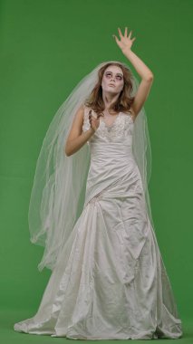 A woman dressed as a dead bride, in a white wedding dress and veil, moves terribly like a zombie. The ghostly bride stands against a green chroma key backdrop for Halloween or spooky projects.