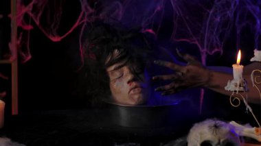 Scary zombie hands place a bowl with a head on the table. A dark room with burning candles, a skull on the table, and creepy cobwebs. Suitable for Halloween themes and holiday media publications.