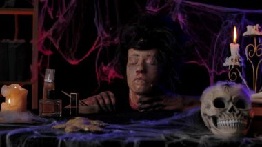 Scary zombie hands apply makeup to a severed dead head. A dark room with burning candles, a skull on the table, and creepy cobwebs. Suitable for Halloween themes and holiday media publications.