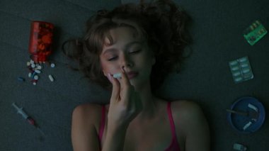 Top view of a young vicious woman in a pink bra putting a pill in her mouth. Scattered around her are pills, blisters, a syringe, and cigarettes, symbolizing addiction and a destructive lifestyle