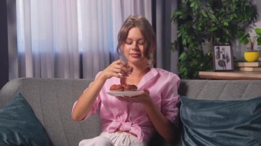 A woman eats mini muffins. A young woman in a pink shirt relaxes on the sofa at home, enjoying a sweet dessert.