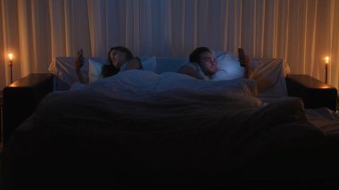 A young man and woman lie in bed, both glued to their smartphones instead of chatting. The theme is gadget and social media addiction, modern relationship problems, and digital addiction at night.