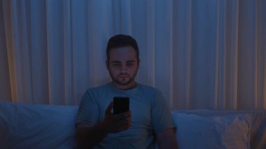 A young man sits in bed at night, illuminated by the light of his smartphone screen. Social media addiction, insomnia, and digital obsession.