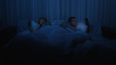 A young man and woman lie in bed, both glued to their smartphones instead of chatting. The theme is gadget and social media addiction, modern relationship problems, and digital addiction at night.