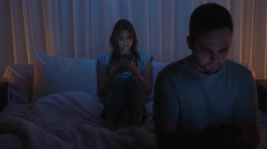 A young man and woman lie in bed, both glued to their smartphones instead of chatting. The theme is gadget and social media addiction, modern relationship problems, and digital addiction at night.