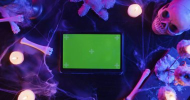 Kafatası, yanan mumlar, örümcek ağı ve yeşil renkli anahtar ekranlı tablet olan bir masanın üst görüntüsü. Cadılar Bayramı teması veya korkunç medya içeriği için reklam veya tanıtım arkaplanı.