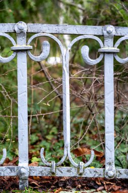 Eski, yıpranmış demir bir çitin yakın çekimi. Çit solgun, açık gri renkte boyanmış, pürüzlü ve dokulu bir yüzeyi ortaya çıkarıyor. Süslü, kıvrımlı demircilik merkezi bir odak noktasıdır..