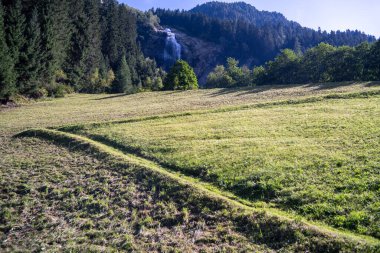Bereketli bir dağ vadisi manzaralı bir çayır ve çarpıcı bir şelalenin nefes kesici geniş bir görüntüsü. Önplanda, yeşil ve sarı otların arasından geçen yol benzeri bir depresyon, gözü yoğun ormana yönlendiriyor..
