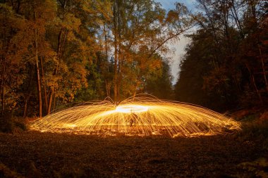 Büyüleyici uzun pozlu bir fotoğraf gece bir ormanda nefes kesici bir ışık resmi performansı yakalar. Parlak altın kıvılcımlardan oluşan bir kubbe, dönen çelik yünün oluşturduğu, merkezi bir noktadan dışarıya doğru yayılır, zemini aydınlatır..