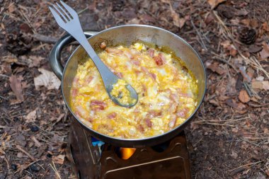 Portatif bir odun ocağında küçük bir kamp tenceresinde pişen lezzetli çırpılmış yumurta ve pastırmalı kahvaltı görüntüsü. Titanyum çatal tencerede duruyor, dişlinin hafif ve pratik doğasını vurguluyor..