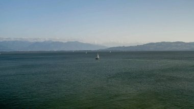 Sakin sular bulutlu bir dağ ufkuna doğru uzanıyor, berrak bir gökyüzünün altında zarif yelkenlilerle benekli. Bu pitoresk manzara engin bir göl ya da körfezin dingin güzelliğini yakalar. Doğa, rahatlama ve açık hava maceraları için mükemmeldir..