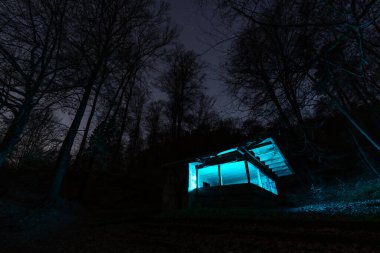 Karanlık ve sessiz bir ormanın derinliklerine kurulmuş, dünya dışı mavi bir ışıkta yıkanmış ahşap bir kulübeyle büyüleyici bir gece sahnesi. Canlı yapay aydınlatma ile çevreleyen doğal karanlık arasındaki çarpıcı tezat..