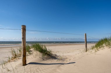 Klasik bir sahil erişim noktasının davetkar bir görüntüsü, ahşap direkler ve yeşil kum tepecikleri tarafından çerçevelenmiş kumlu bir yol, nazikçe parlayan okyanusa doğru gidiyor. Uçsuz bucaksız mavi gökyüzünün altında, kumsalın engin genişliği nazik dalgalarla karşılaşıyor..